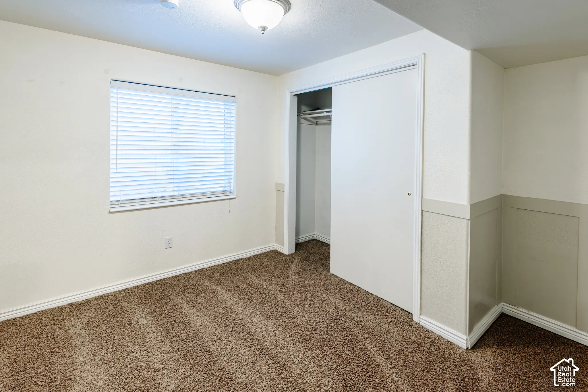 Room featuring neutral wall-to-wall carpeting, wainscoting detail, ceiling-mounted light fixture, window with horizontal blinds, and a built-in closet with sliding doors