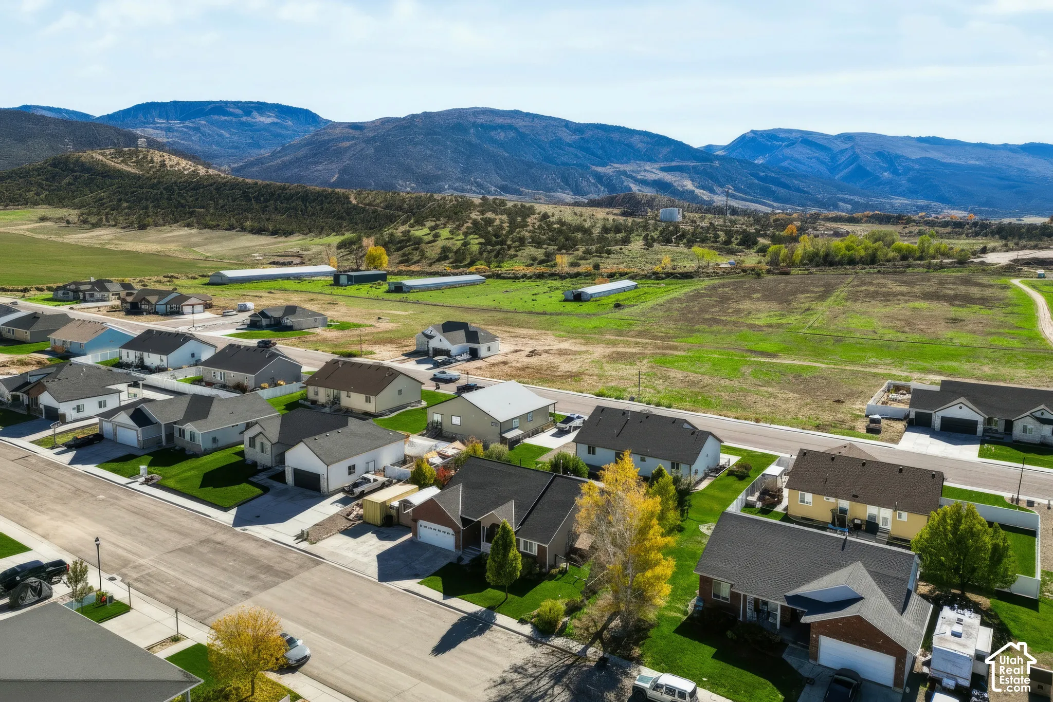 Aerial perspective of a residential neighborhood featuring asphalt paved streets, manicured green lawns, multi-pitched rooflines, and mountain range scenery