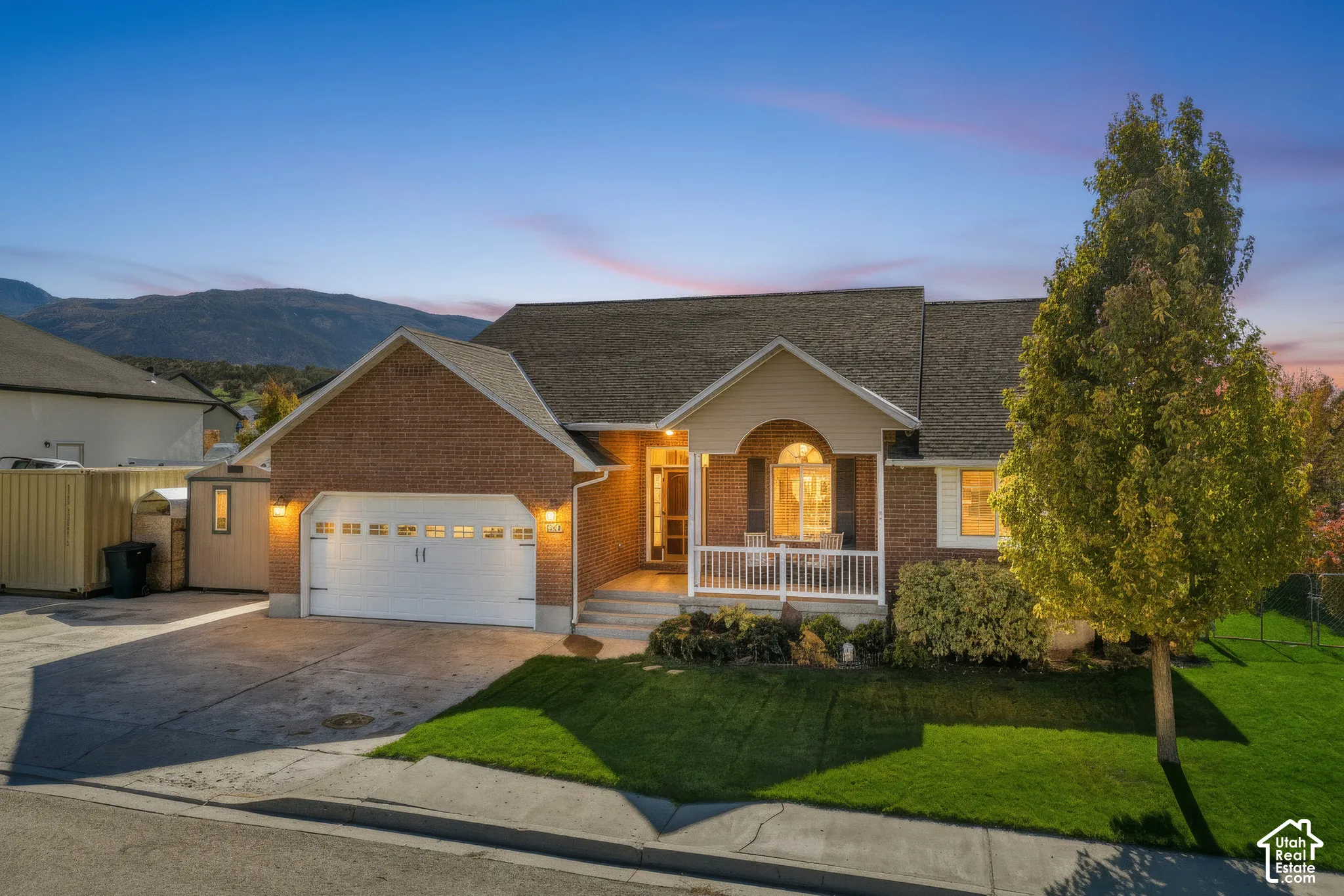 Exterior featuring a brick and siding facade, an arched entry porch with white railings, a two-car garage, and a manicured front lawn set against a mountain backdrop