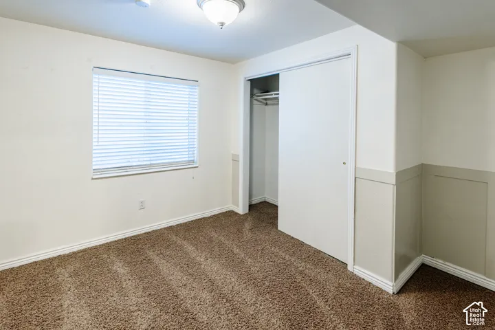 Room featuring neutral wall-to-wall carpeting, wainscoting detail, ceiling-mounted light fixture, window with horizontal blinds, and a built-in closet with sliding doors