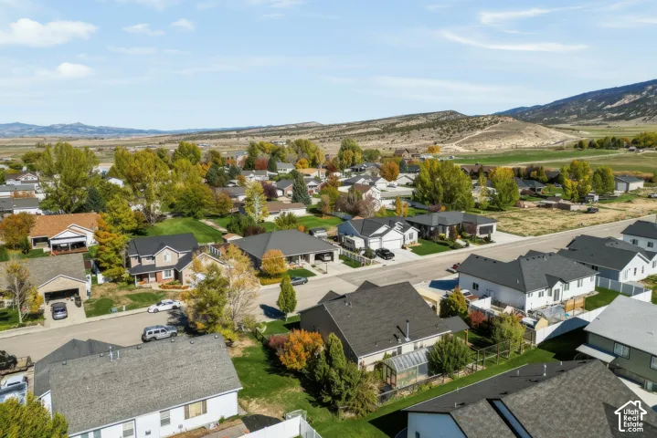 Aerial perspective of a suburban neighborhood featuring paved streets, mature deciduous trees, residential dwellings with pitched roofing, and an expansive mountain range backdrop