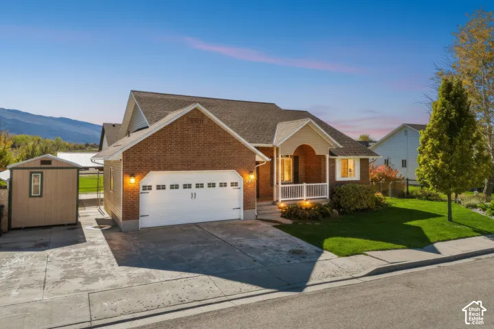 Brick exterior featuring an arched entryway, covered porch with white railing, two-car garage, and detached storage shed, set against a mountain backdrop