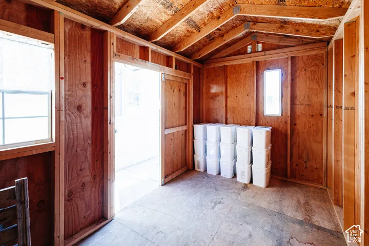 Outbuilding interior featuring exposed wood-stud framing, plywood walls, vaulted roof rafters, two windows, and a wood-panel door