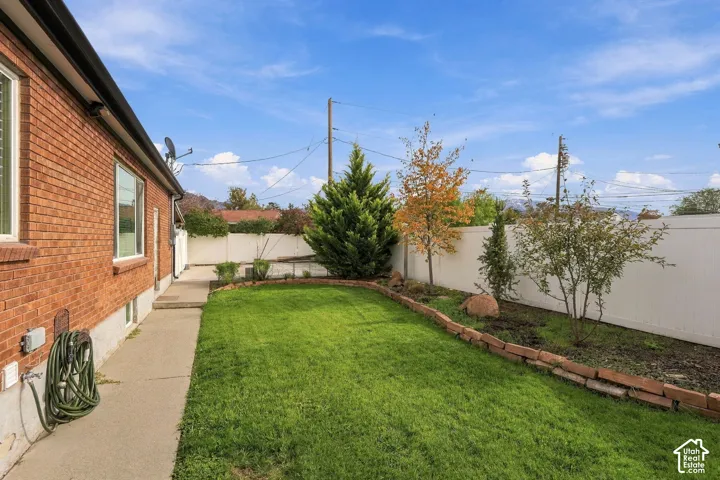 Backyard featuring a red brick exterior, concrete walkway, vinyl fencing, manicured lawn, and stone-bordered garden beds