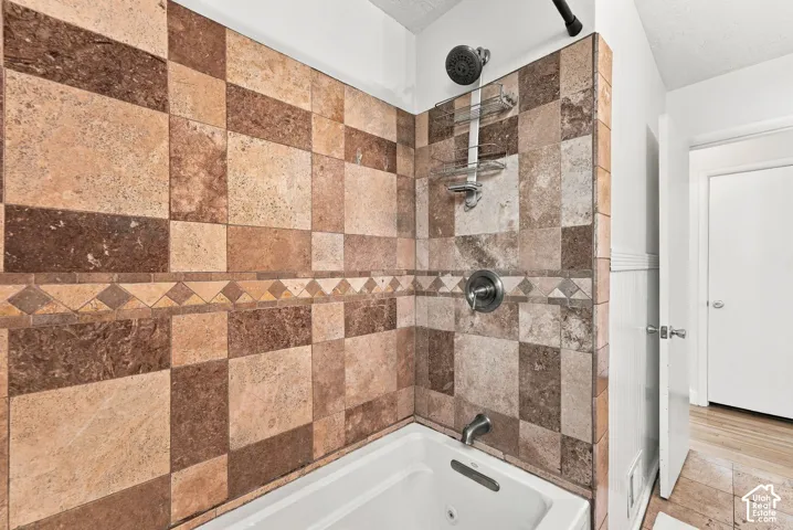 Bathtub enclosure featuring multi-toned stone tile surround, decorative mosaic tile accent border, wall-mounted metal plumbing fixtures, and integrated metal shower caddy