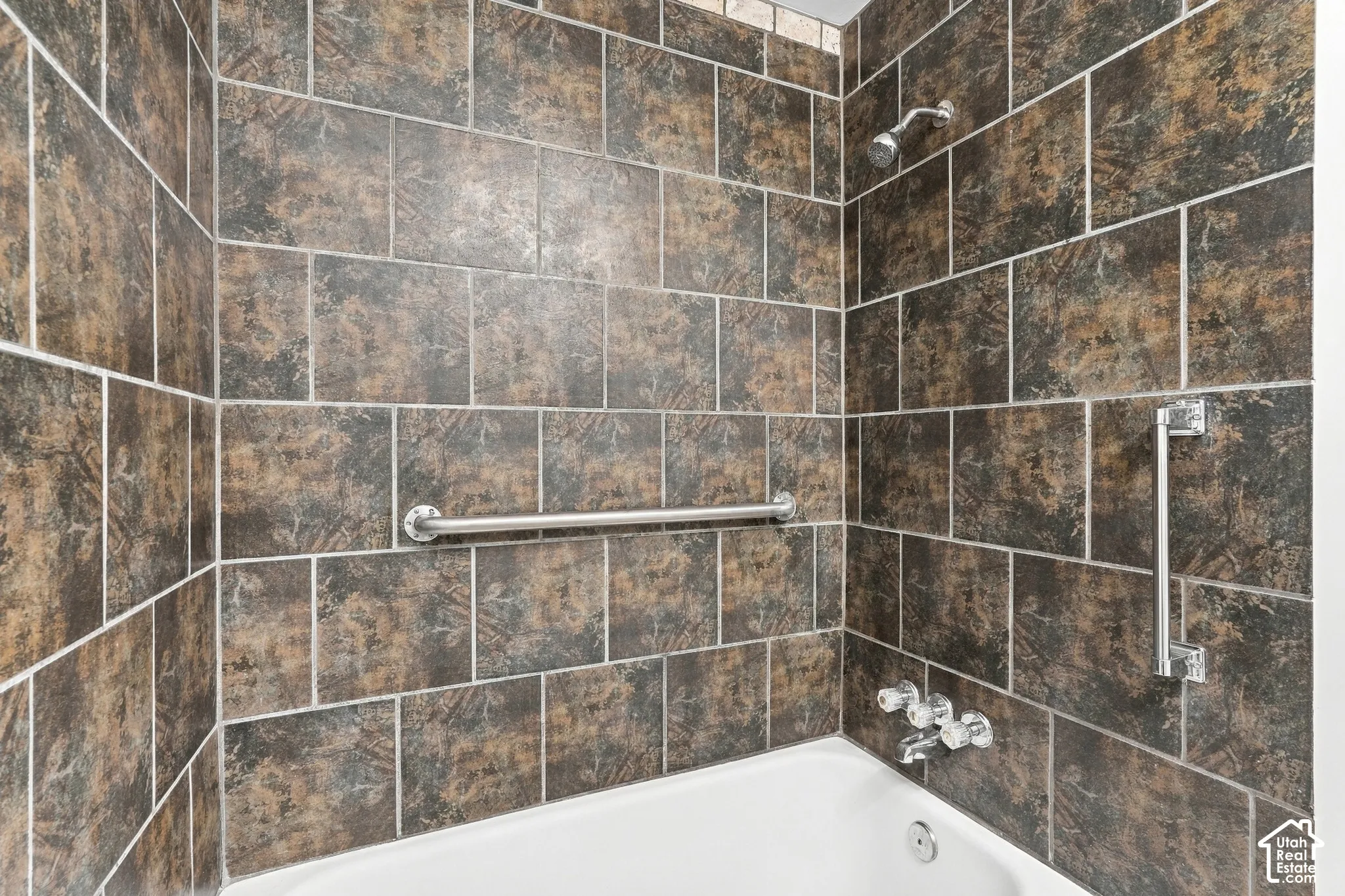 Full-height shower surround featuring multi-tonal, earth-toned square tiles, a white acrylic tub, stainless steel wall-mounted grab bars, and a classic chrome shower head and faucet fixture