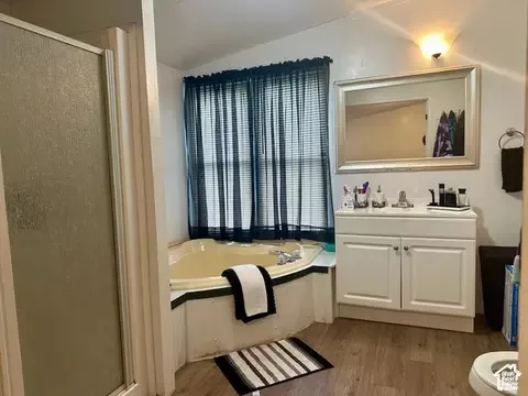 Bathroom featuring a corner soaking tub, separate shower enclosure with a glass door, white vanity cabinet with a integrated countertop sink, and wood-finish flooring
