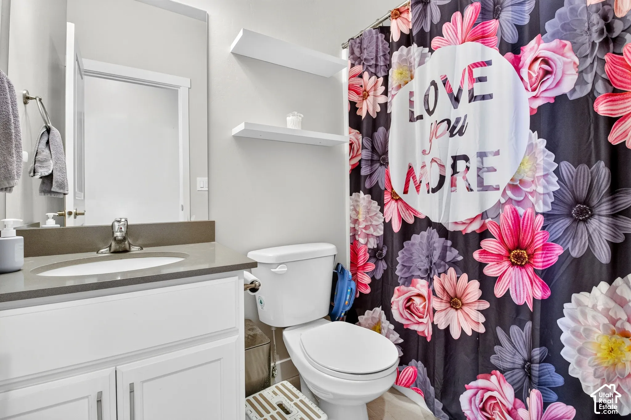 Bathroom vanity featuring white cabinetry, a solid surface countertop with an integrated sink, and polished chrome hardware, complemented by wall-mounted floating shelving and a full-height mirror