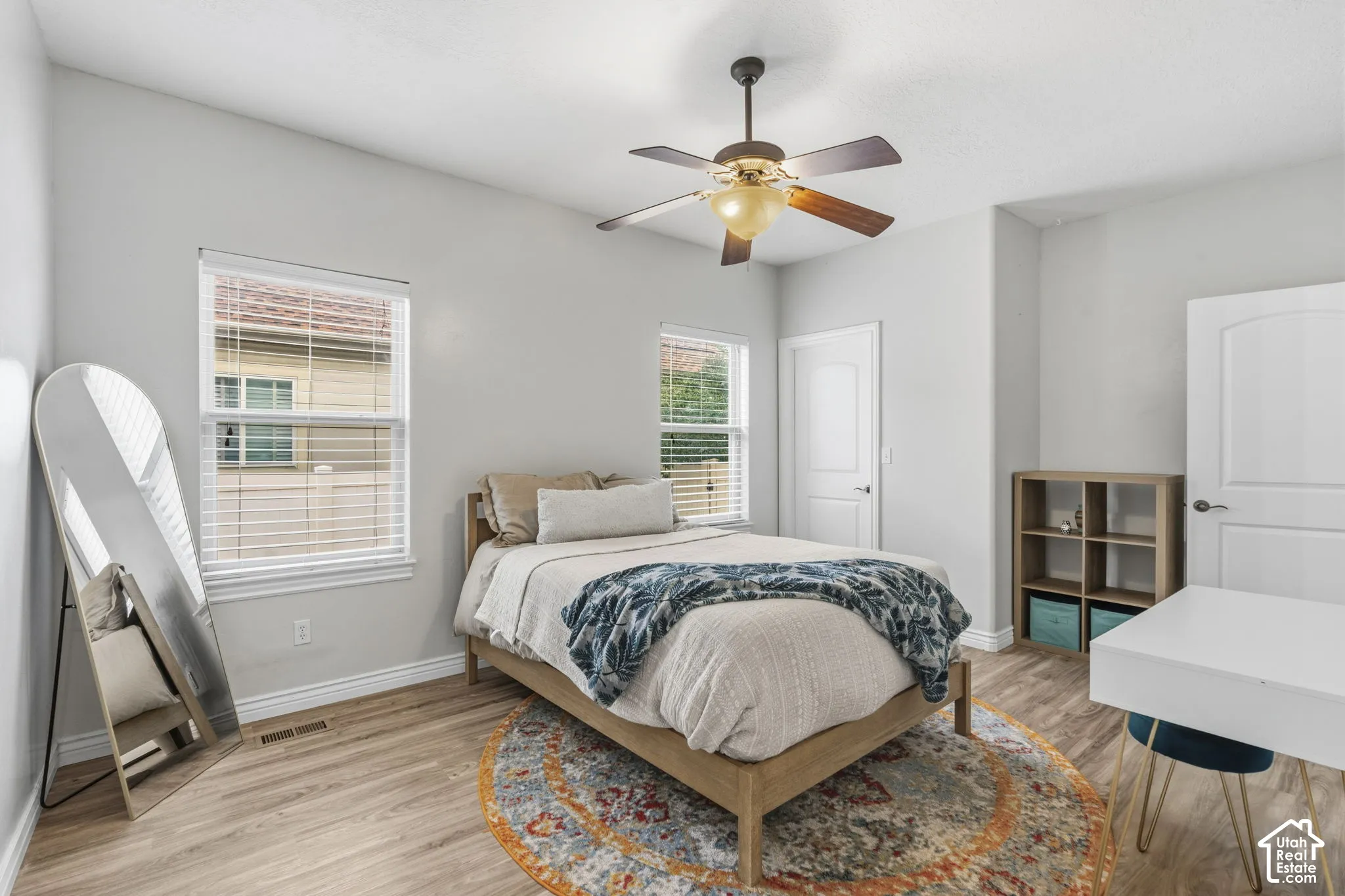Neutral-toned interior featuring wood-finish flooring, dual windows with horizontal blinds, a multi-blade ceiling fan with integrated lighting, and white paneled doors