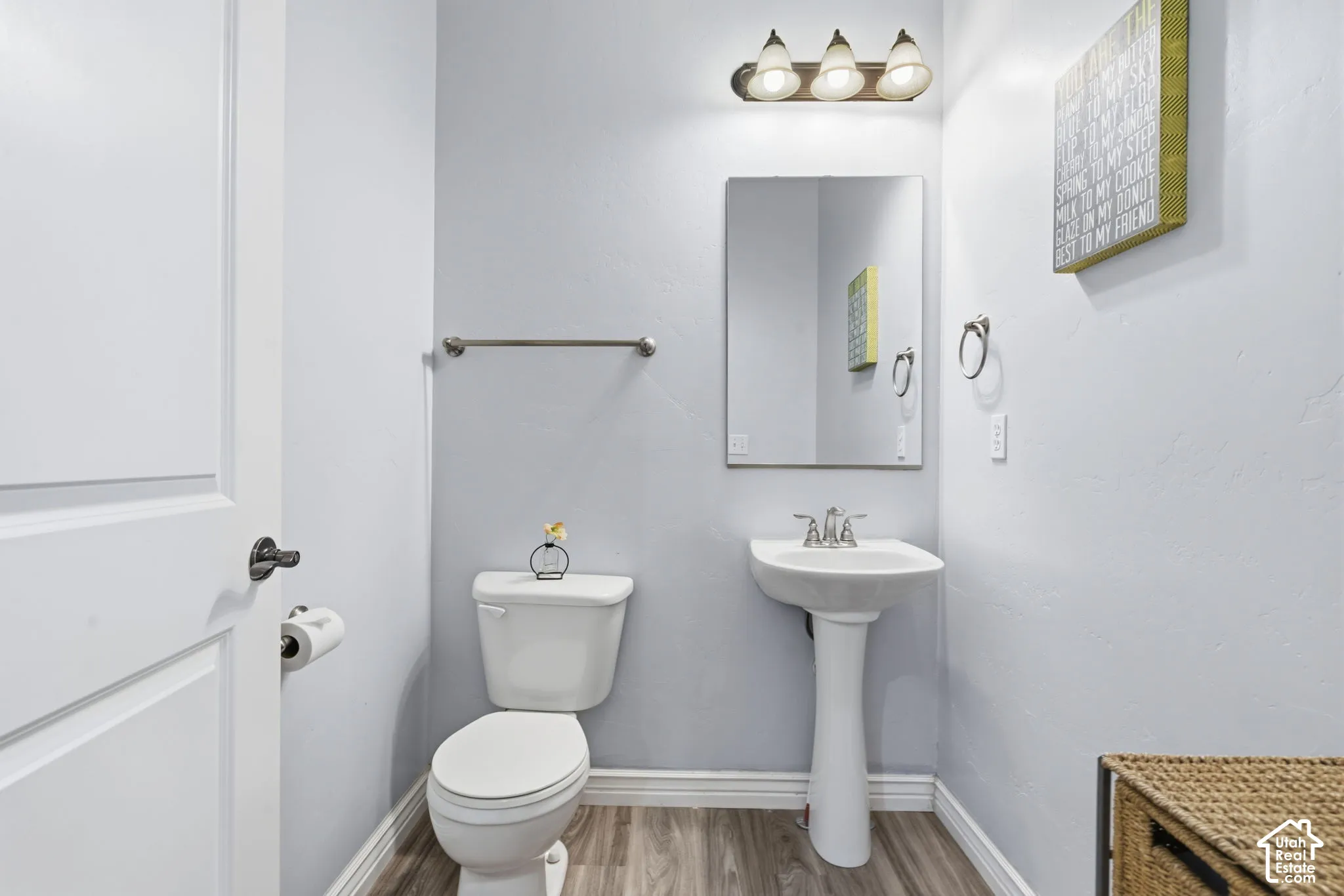 Powder room featuring a white pedestal sink, chrome fixtures, frameless rectangular mirror, wood-finish flooring, and a three-bulb vanity light