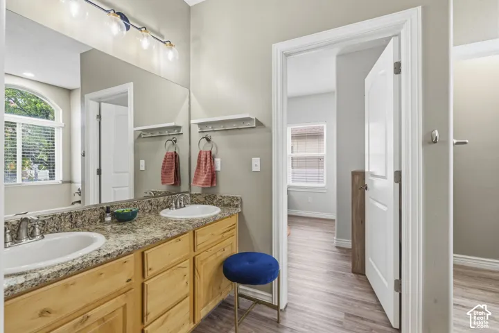 Dual-sink vanity with stone countertop and light wood cabinetry, large vanity mirror, arched window with blind, and wood-finish flooring