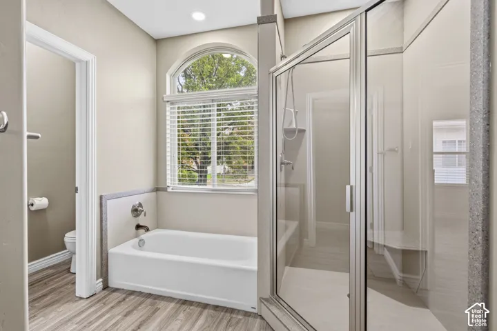 Soaking tub with a tiled surround, arched window, enclosed glass shower, wood-finish flooring, and recessed ceiling lighting