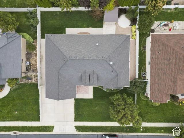Aerial perspective of a property featuring an expansive lawn, concrete driveway, fenced perimeter, shingle roof, and mature landscaping