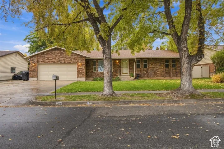 Single-story exterior featuring a brick facade, attached two-car garage, concrete driveway, and mature trees