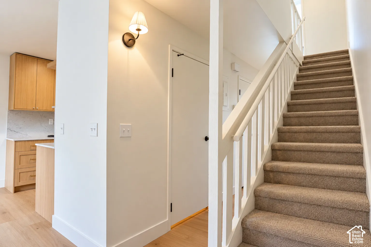 Carpeted staircase with white railings, wood-finish flooring, light-toned cabinetry, and wall-mounted sconce lighting