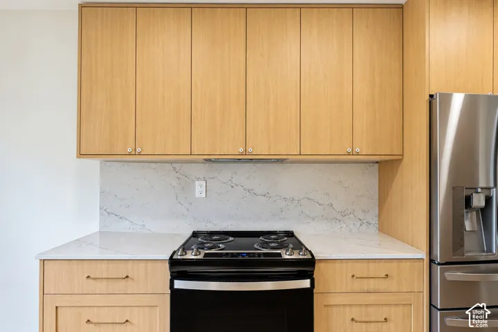 Kitchen featuring light-toned cabinetry, stone-finish countertops, full-height stone-finish backsplash, stainless steel refrigerator, and integrated black range