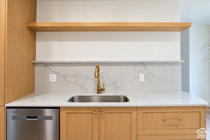 Light-wood cabinetry, stone-surface countertop, full-height stone backsplash with matching ledge, integrated stainless steel dishwasher, and floating wood shelf with brass-finish faucet hardware
