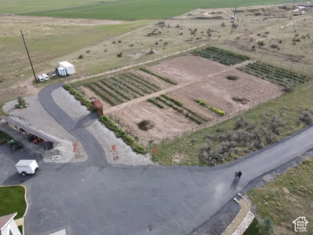 Aerial view of a sprawling lot featuring an expansive cultivated garden area, fenced perimeter, paved driveway, and open-sided storage structure