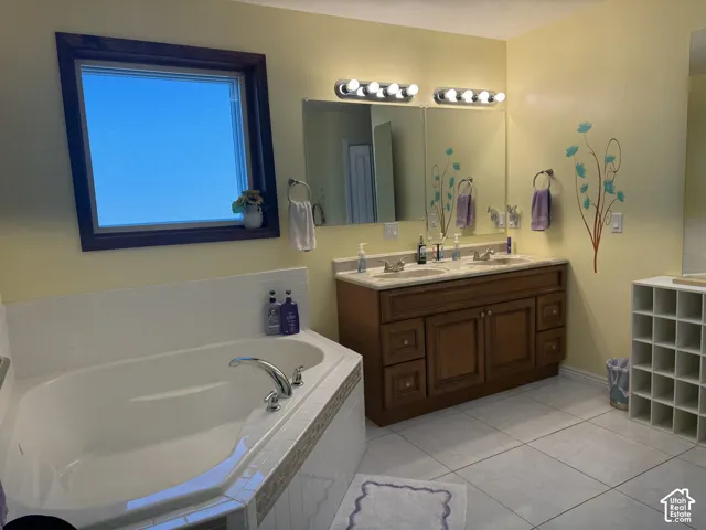 Primary bathroom featuring a soaking tub with tile surround, double-sink vanity with stone countertop, large wall mirrors, and tile flooring