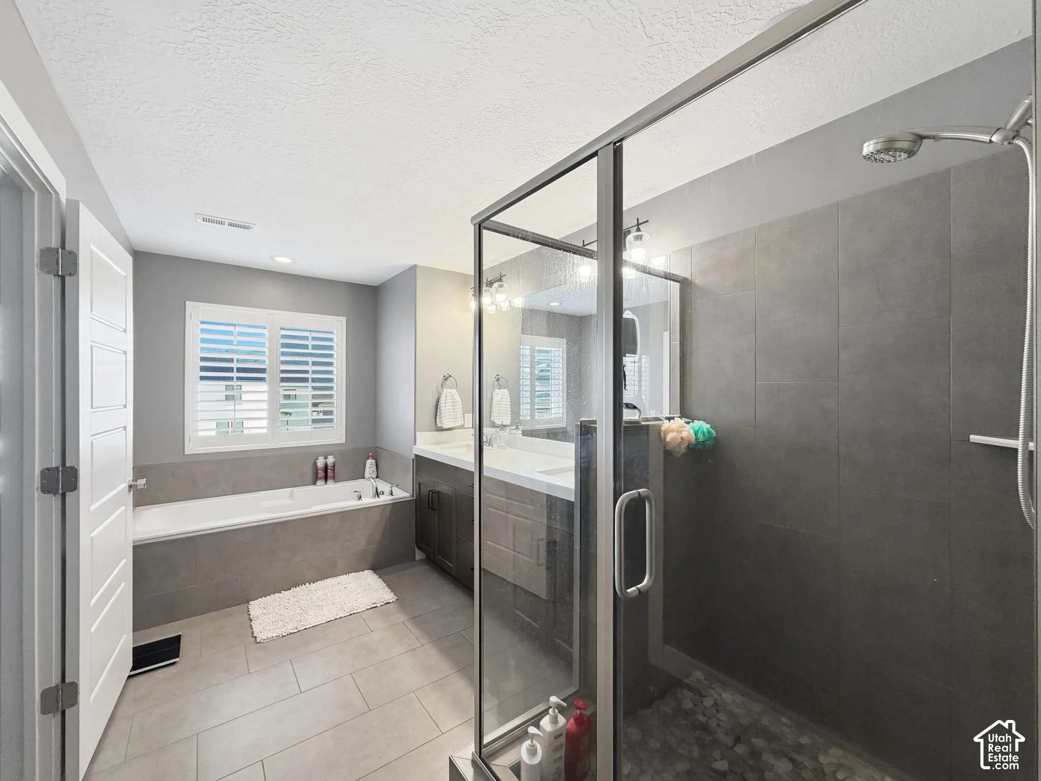Bathroom featuring a walk-in shower with large-format tile surround, deep soaking tub with tile deck, dual-sink vanity with dark cabinetry, and plantation shutters