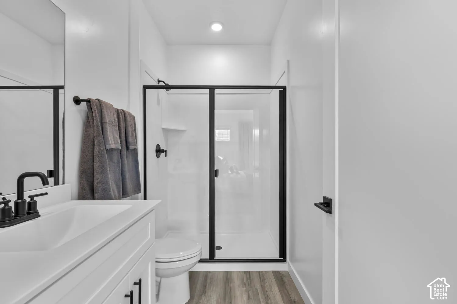 Bathroom featuring a white vanity with a composite countertop, matte black fixtures, wood-finish flooring, and a walk-in shower with a black-framed glass enclosure
