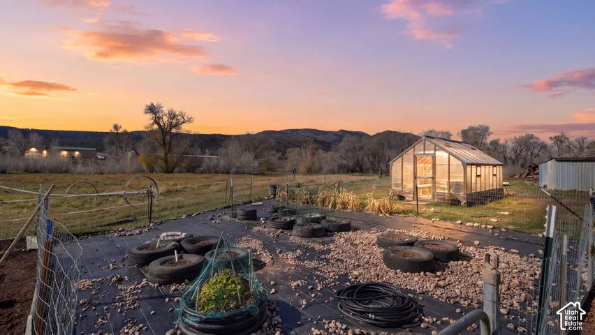 Outdoor garden space featuring a glass-paneled greenhouse, woven wire perimeter fencing, and stone-mulched planting areas with mountain backdrop