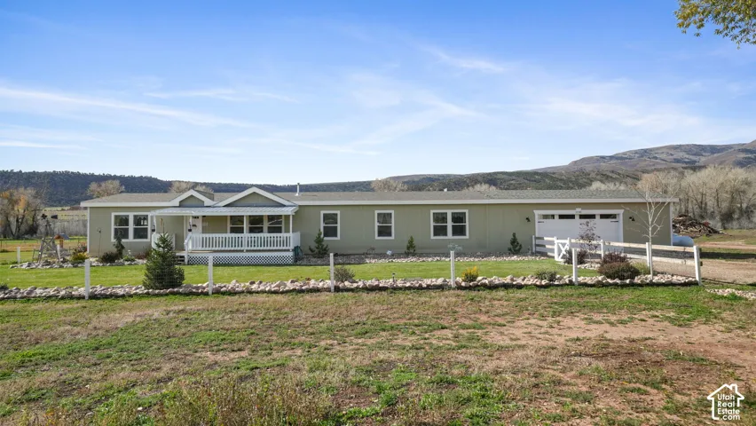 Single-level exterior featuring horizontal siding, a covered front porch with white railing, an attached garage, and a rock-bordered front lawn set against a mountain backdrop