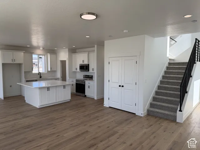 Kitchen featuring white shaker cabinetry, quartz countertops, a central island, stainless steel appliances, and wood-finish flooring with an integrated staircase