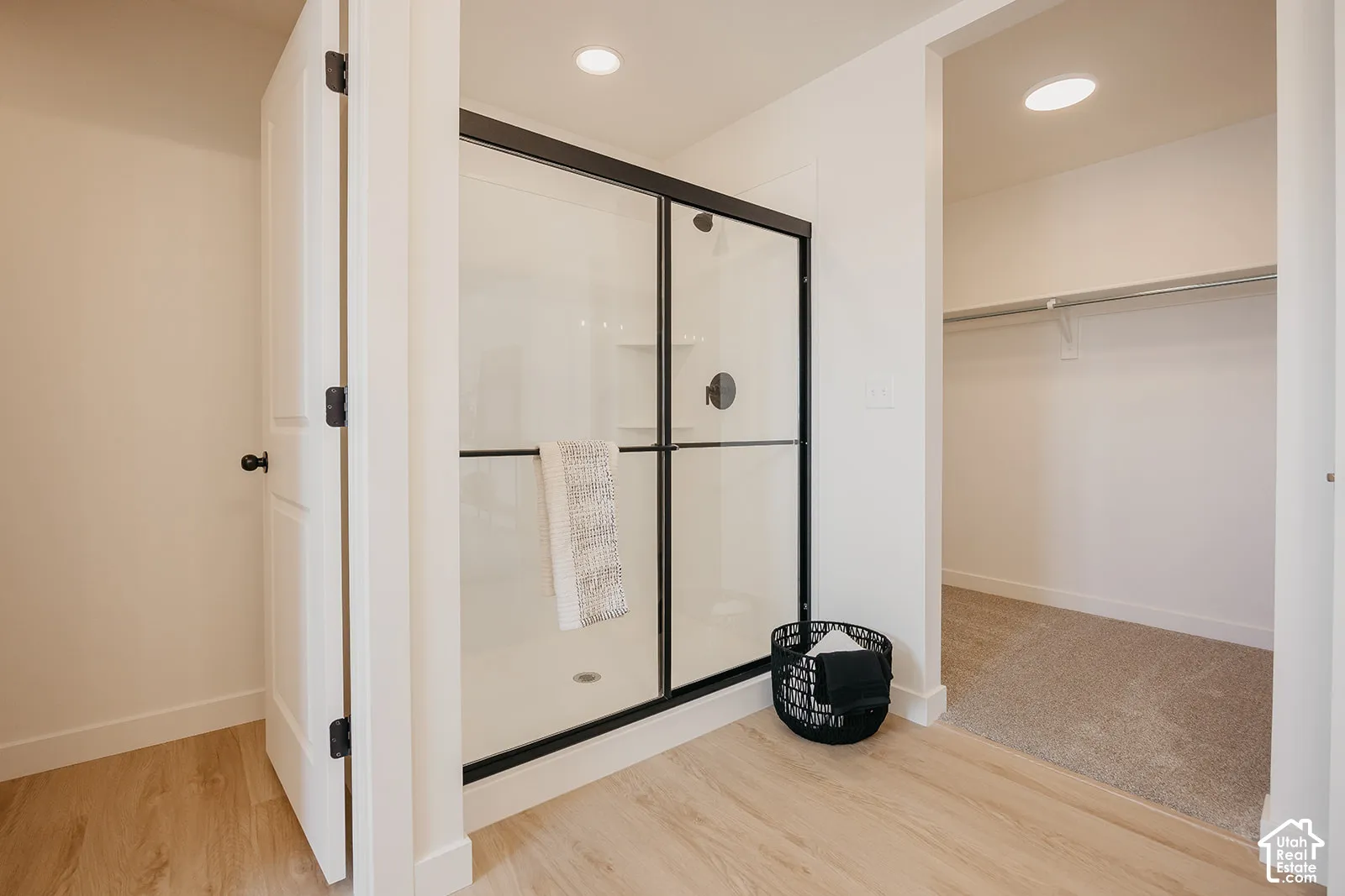 Walk-in shower featuring a glass enclosure with a black frame, recessed lighting, and wood-finish flooring, accompanied by a separate storage area with installed shelving and carpeting