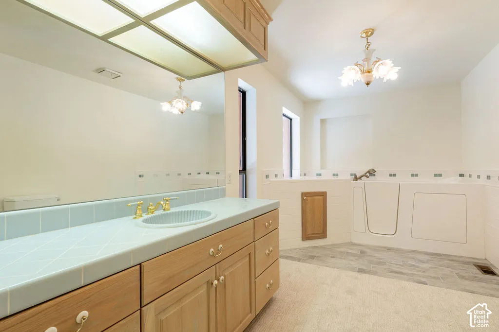 Second bedroom bathroom featuring a vanity with wood-finish cabinetry, a tile countertop with an integrated basin, a large wall-mounted mirror with integrated lighting, a multi-fixture chandelier, and a walk-in tub with tile surround