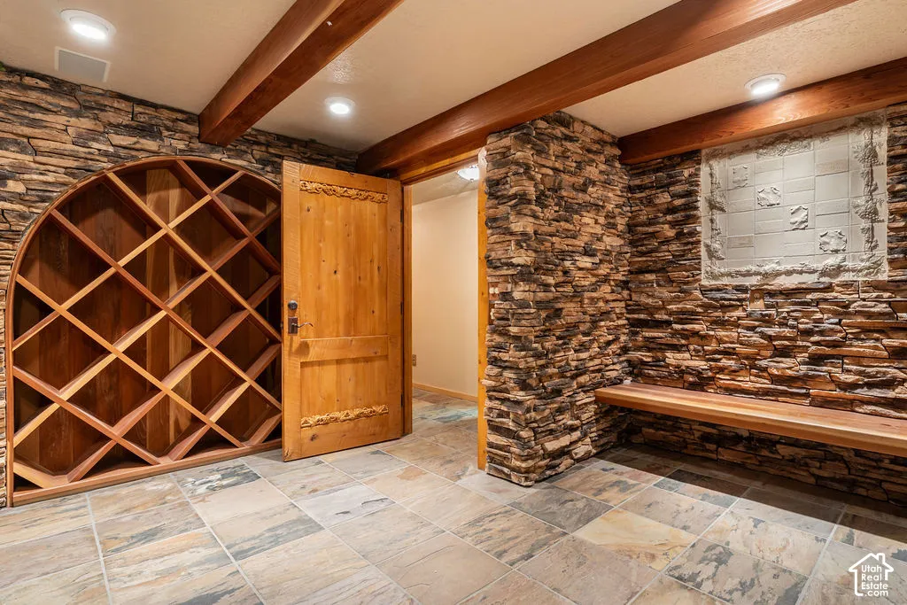Stacked stone wall accents, built-in arched wooden wine shelving, exposed timber ceiling beams, natural stone tile flooring, and a floating wood bench