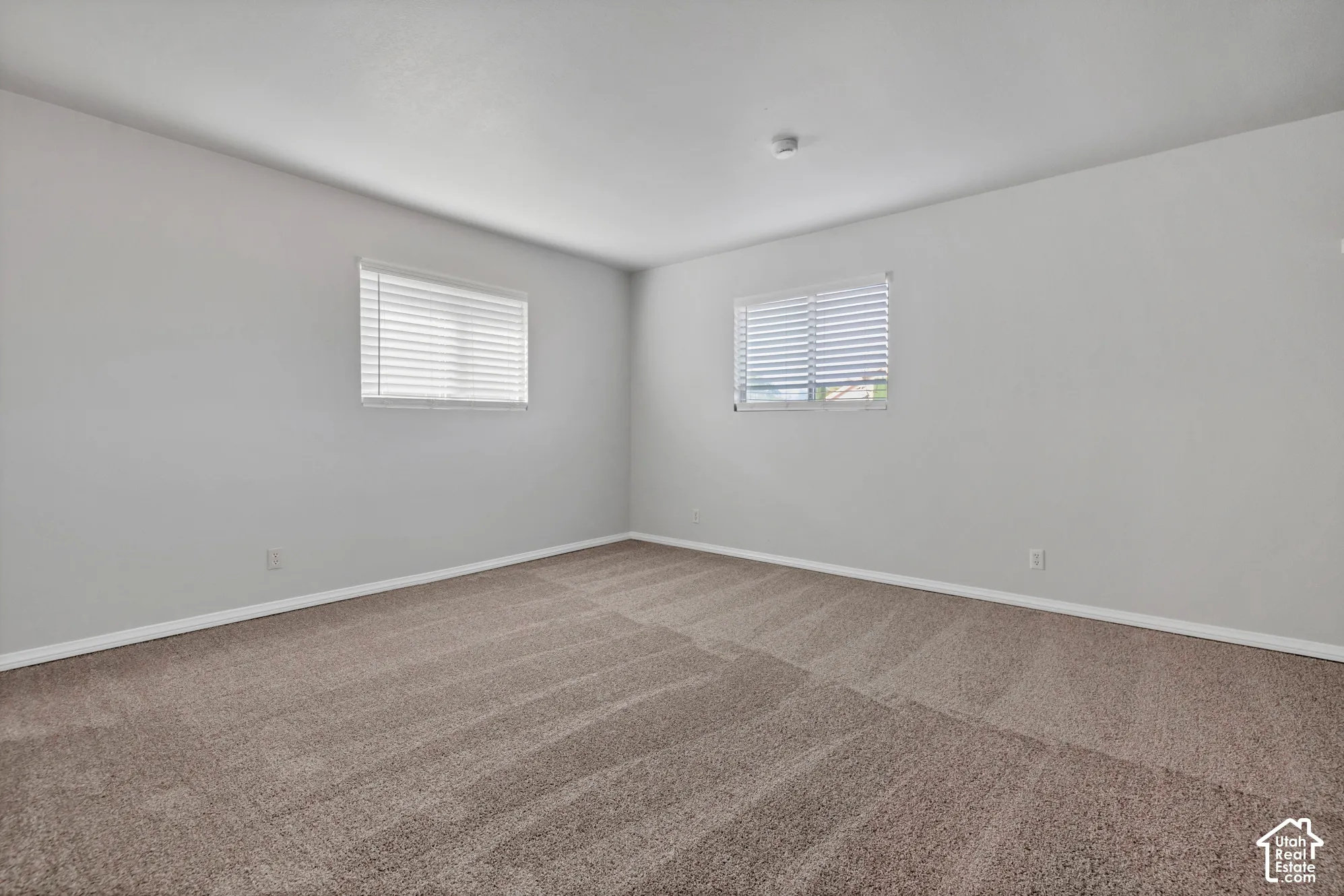 Neutral-toned carpeted flooring, two windows with horizontal blinds, and smooth-finish wall surfaces