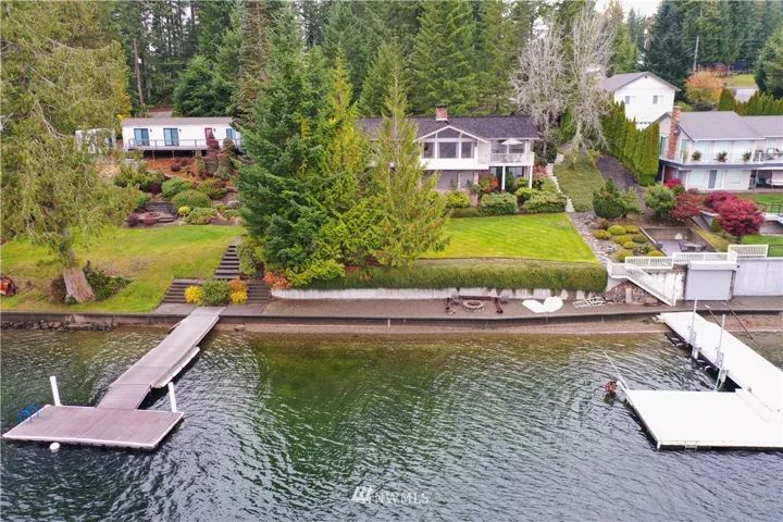 View of the property from the water, the double lot runs from where the tall trees start by the shared steps on the left to the end of the white fence on the right.  View includes the dock on the right, boat house just to the right of the dock , the bulkhead with fire pit area, and beautifully landscaped yard!