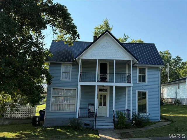 View of front facade featuring a porch, a balcony, a chimney, a front lawn, and a metal roof