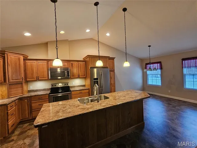 Kitchen with brown cabinets, appliances with stainless steel finishes, light stone counters, pendant lighting, and a center island with sink