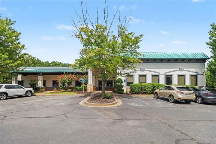 Single-story commercial building featuring a green metal roof, a stucco exterior, multiple window openings, and an asphalt parking lot