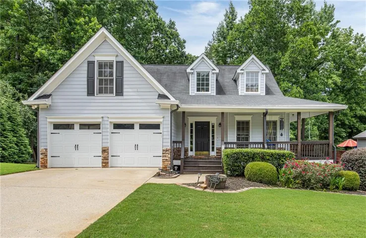 Light blue siding exterior featuring a covered front porch with dark wood columns and railing, two dormer windows, and a two-car garage with stone accents