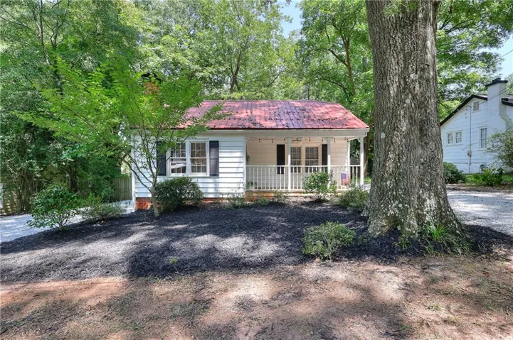 Charming cottage featuring a wraparound porch with decorative railings, white horizontal siding, and a distinctive pitched roof with metal sheathing
