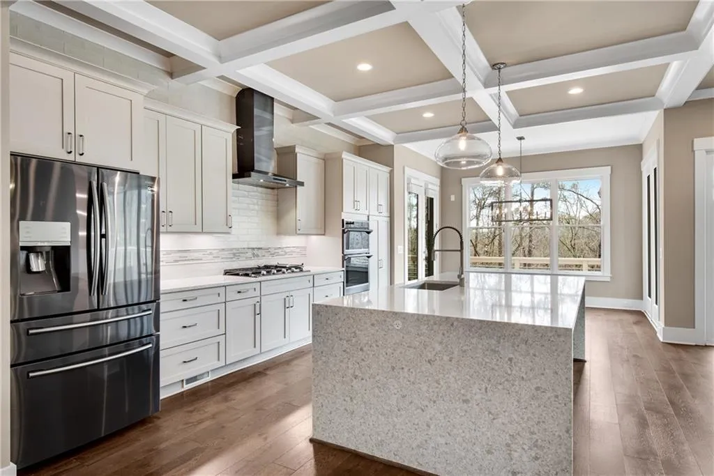Kitchen featuring stainless steel appliances, light stone counters, coffered ceiling, a kitchen island with sink, and decorative backsplash