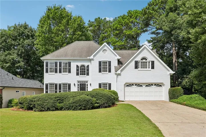 Two-story white brick residence featuring a prominent front entrance with sidelights, a two-car garage, mature landscaping, and a concrete driveway