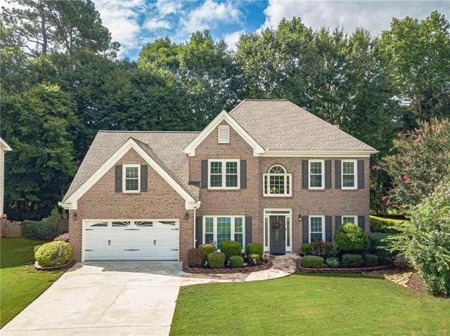 Traditional brick exterior featuring a two-car garage, mature landscaping, a stone pathway, and multiple windows with dark shutters