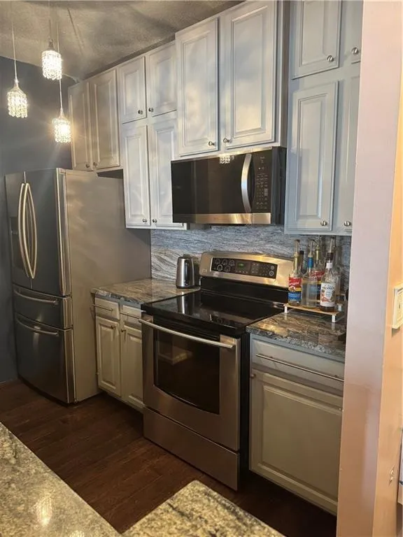 Kitchen featuring appliances with stainless steel finishes, backsplash, dark stone countertops, hanging light fixtures, and dark wood-style floors