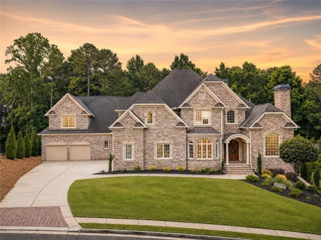 Stone façade residence featuring multiple gables, a covered entryway, a three-car garage, manicured lawn, and mature trees