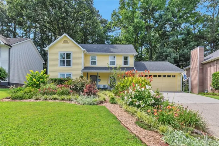 Yellow siding exterior featuring a covered porch, attached two-car garage, manicured lawn, and mature landscaping