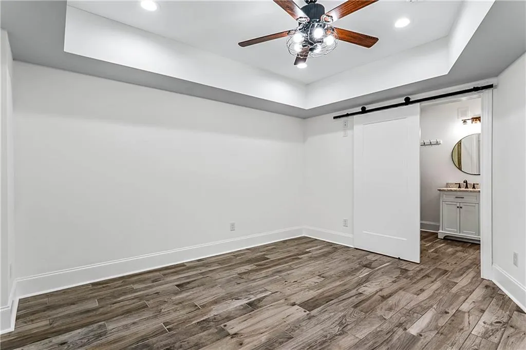 Unfurnished bedroom with a barn door, a tray ceiling, a ceiling fan, wood finished floors, and recessed lighting