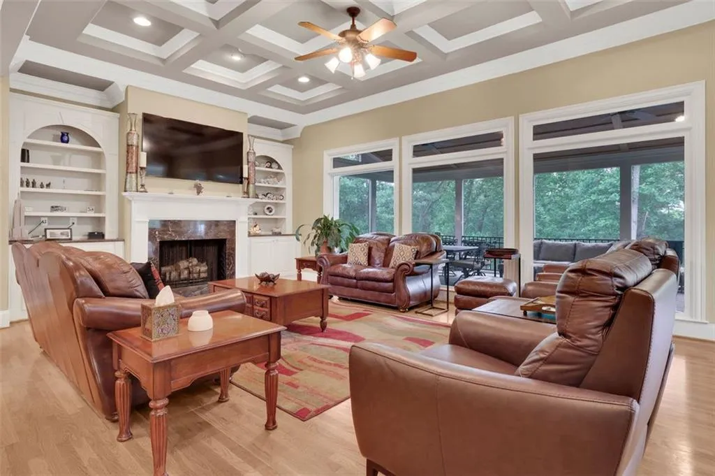 Family room featuring large windows overlooking screened porch and tree tops, ceiling fan, a fireplace, coffered ceiling, and light wood-type flooring