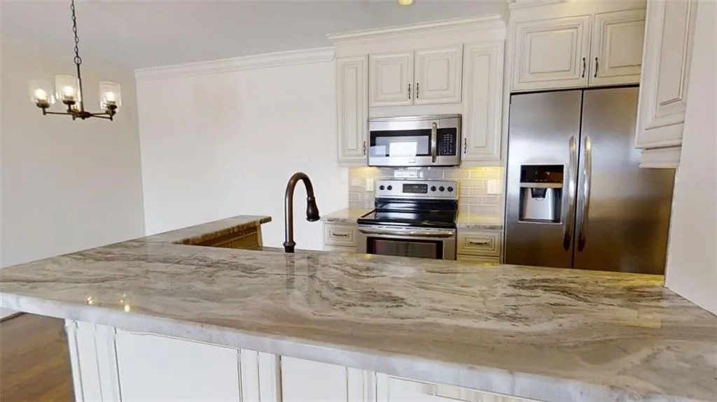 Kitchen with stainless steel appliances, light stone counters, hanging light fixtures, white cabinetry, and crown molding