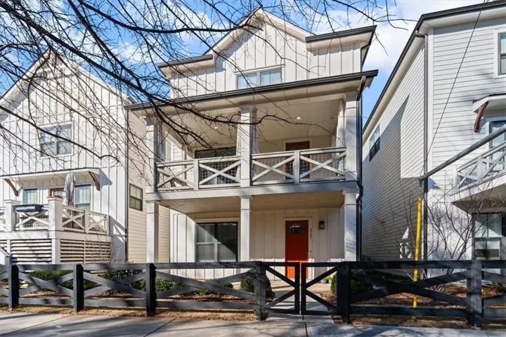 Modern farmhouse exterior featuring vertical board and batten siding, a prominent second-story balcony with X-pattern railings, a contrasting red entry door, an additional lower-level porch, and a black wooden fence with a matching gate