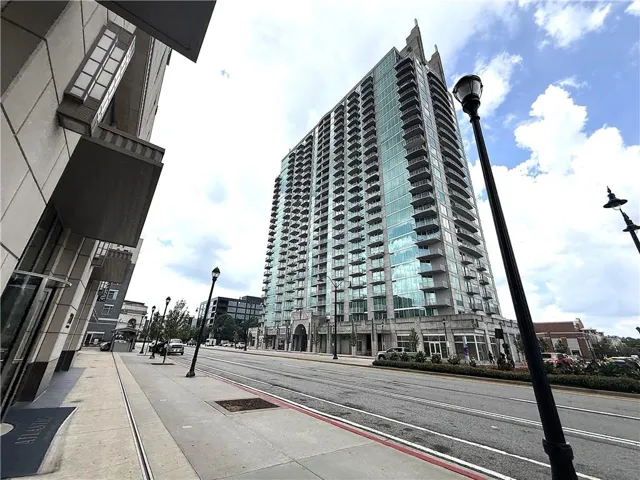 Modern high-rise residential building featuring extensive glass facade, multiple private balconies, and a prominent street-level entrance