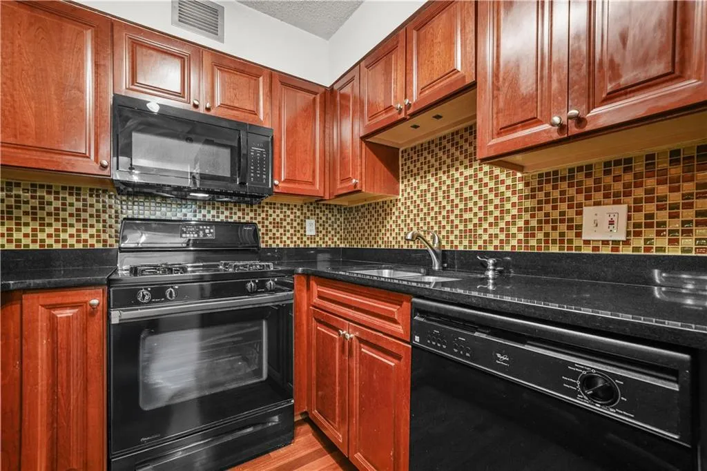 Kitchen with black appliances, backsplash, dark stone counters, brown cabinetry, and wood finished floors